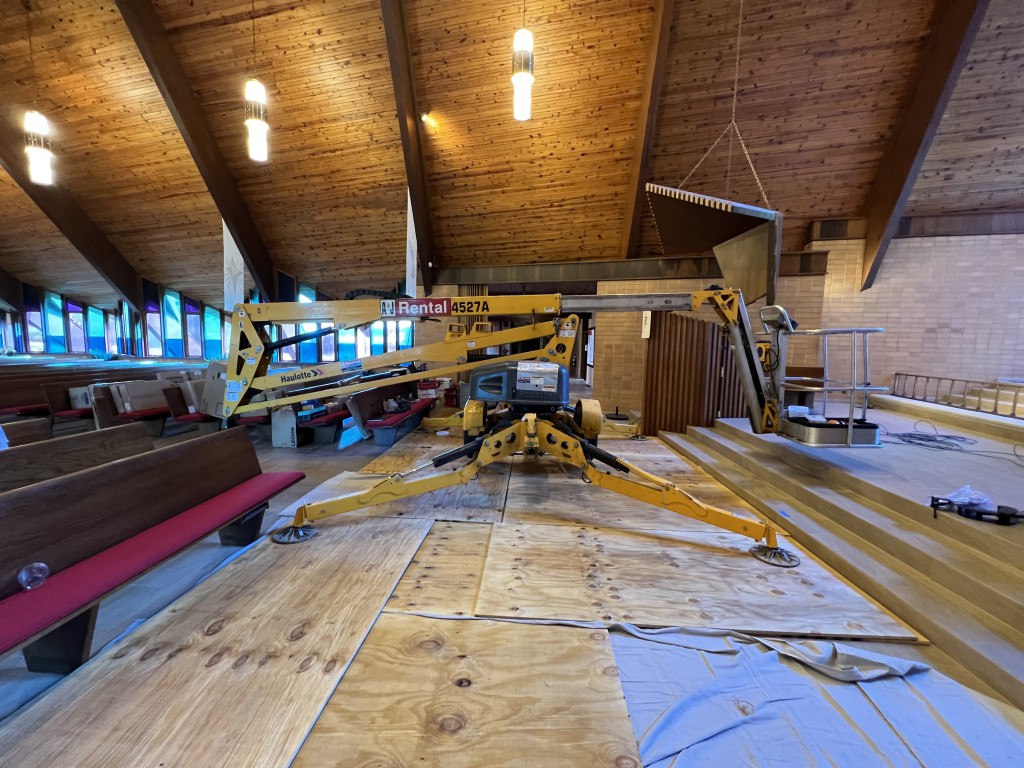 An articulating lift, a heavy-duty piece of equipment designed to reach high spaces, sits on a temporary plywood floor in a church sanctuary.