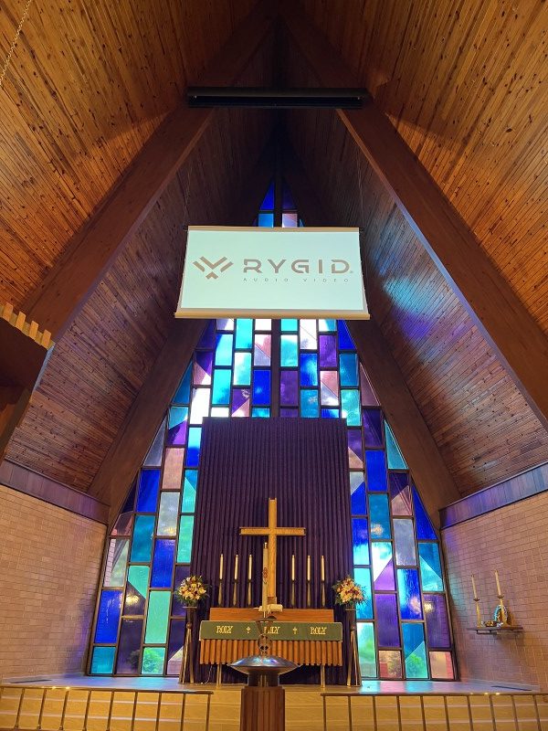 A digital projection screen with the words "RYGID AV" hangs from the ceiling of a beautiful church sanctuary with blue and purple stained-glass windows behind it.
