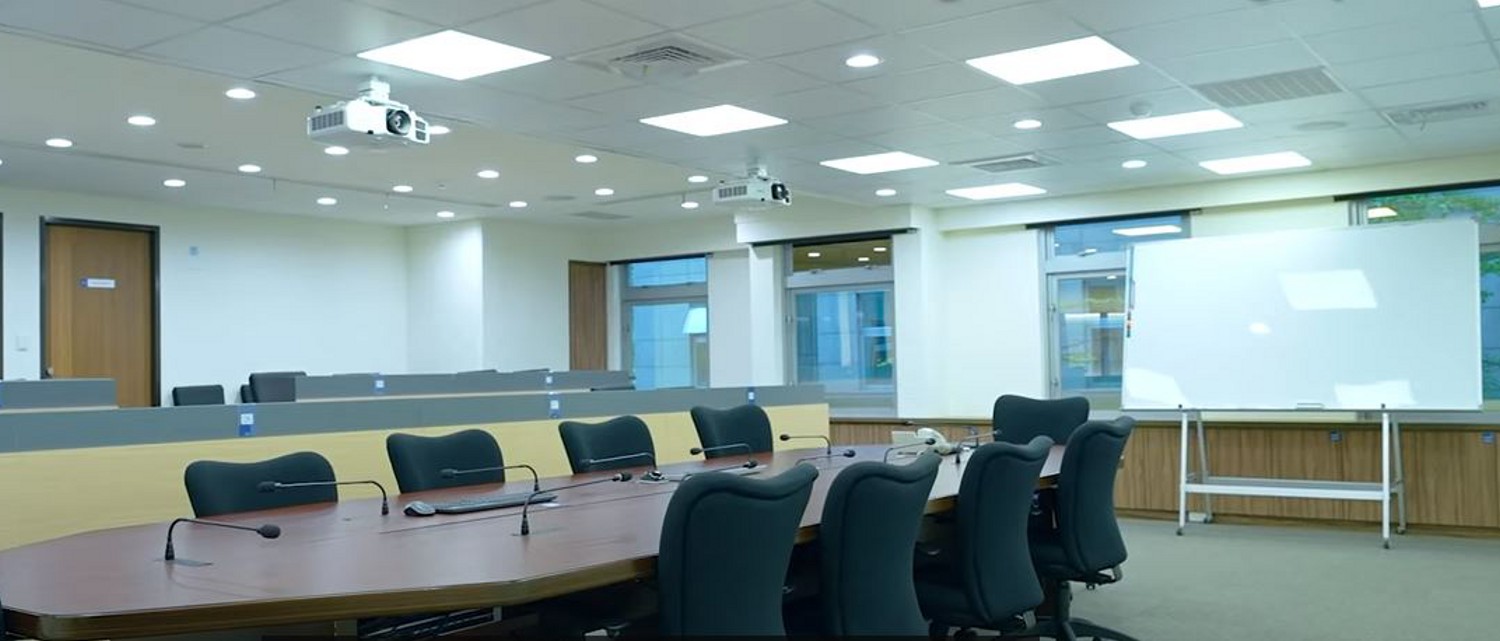 A sleek, modern conference room with ceiling-mounted projectors and wired microphones around an oval conference table. A glossy dry-erase board sits behind them.