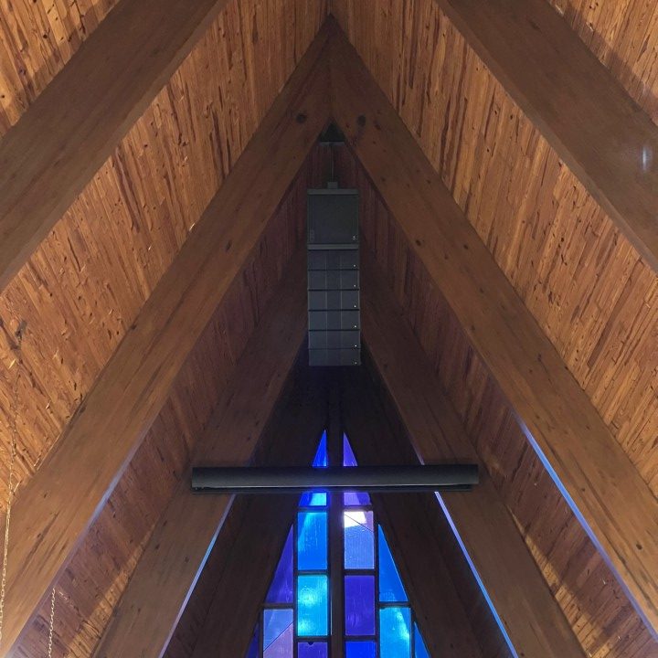 A tall, peaked roof in a church, lined with beautiful wood, with a speaker and mounted projection screen in front of a stained glass windows.
