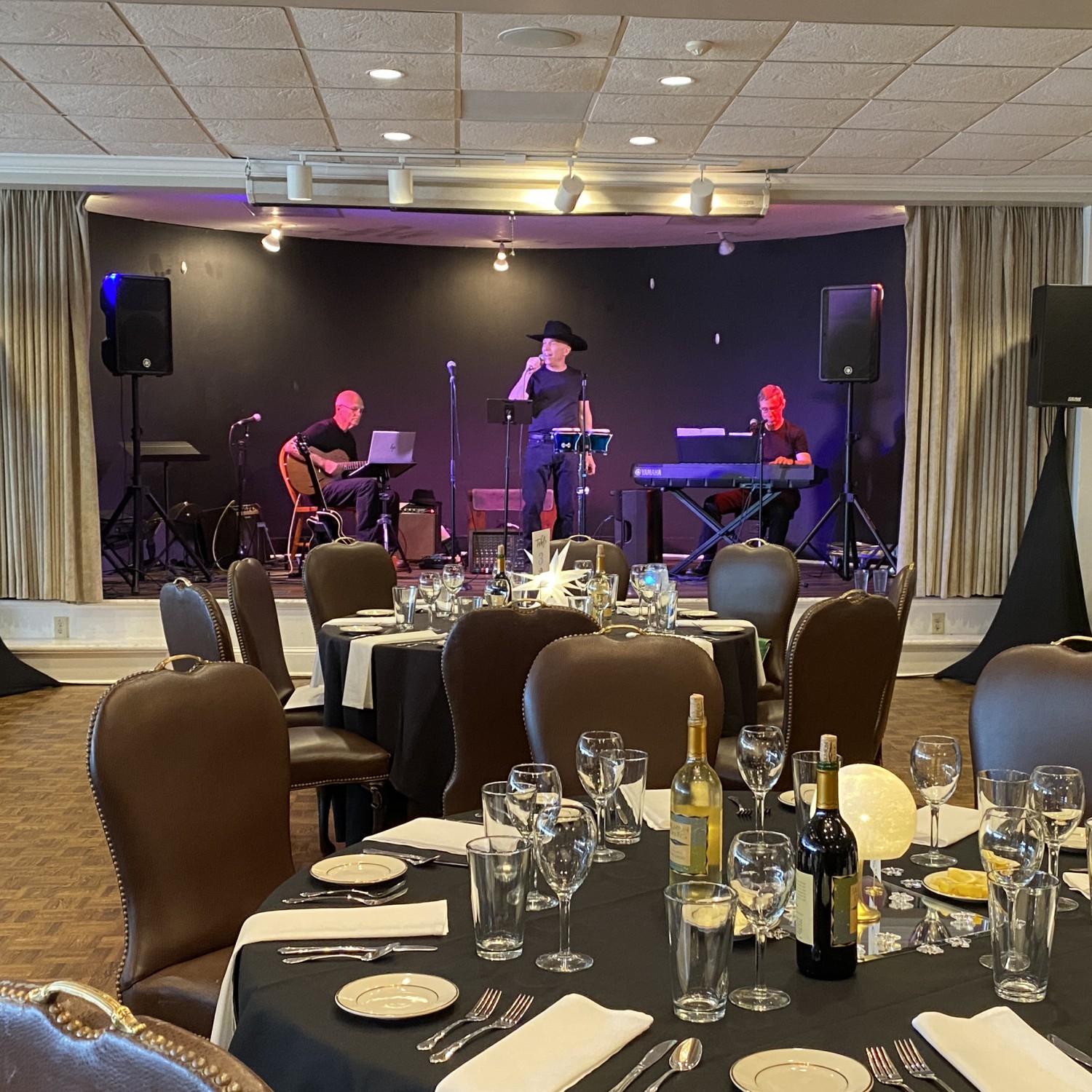 An elegantly decorated round dining table with wine bottles and empty glassware and plates sits in the foreground; a stage with a signer in a cowboy hat and jeans, an electronic keyboard and player, and a guitar player, do a sound check for the event that is happening soon.