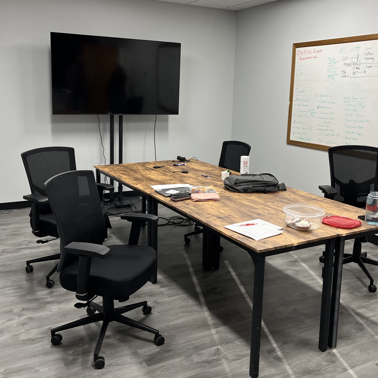 A wooden-topped table surrounded by four office chairs on wheels. Papers and writing instruments clutter the table. A dry erase board with notes on it and a large, flat-screen TV hang on the wall.