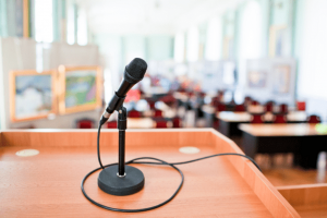 In a bright auditorium, a microphone sits on a wooden podium with a blurred-out background. A commercial audio visual system helps everyone in the audience see and hear what is happening on stage.