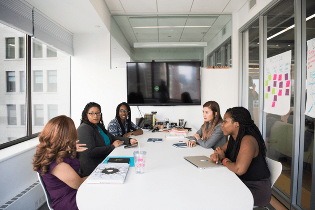 Five women sit in a glass huddle room, brainstorming ideas for work. A large TV screen hangs on the wall behind them, displaying ideas they come up with.