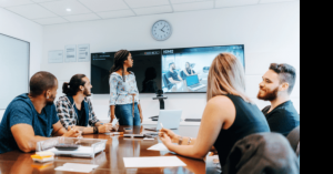 Five coworkers sit around a table in a conference room; a camera captures their image and displays it on a large screen behind them and to virtual attendees. The AV tech makes the meeting room work better for both their local and remote employees.