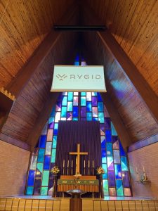 A digital projection screen with the words "RYGID AV" hangs from the ceiling of a beautiful church sanctuary with blue and purple stained-glass windows behind it.