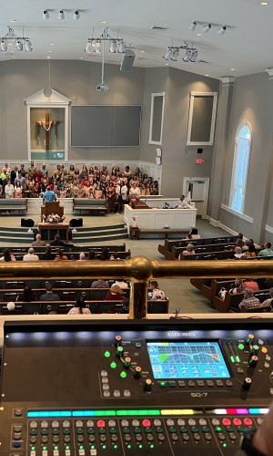 A photo from a balcony; most of the image captures a live church session with a chorus and worshippers in the pews. In the foreground, a church soundboard is lit up, controlling and mixing the sound.