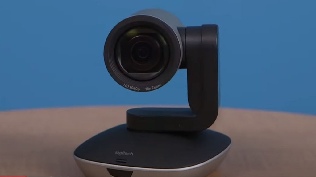 A Logitech HD 1080p 10x Zoom camera sitting on a wooden conference room table in front of a bright blue wall.