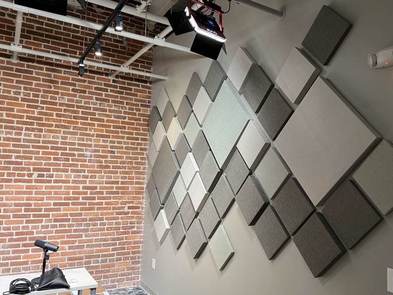 A pattern of grey and white square acoustical wall panels hang next to an exposed brick wall in a podcast recording studio.