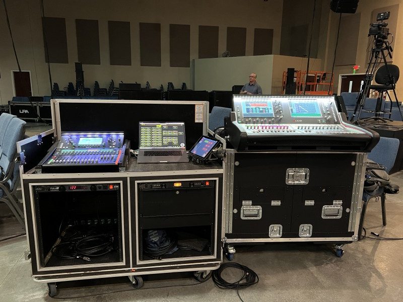 A collection of four different types of soundboards sit on the boxes used to transport the equipment, as they are being set up for a complex live event.