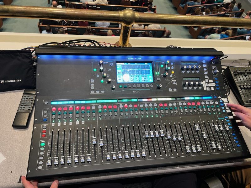 A complete picture of an Allen & Heath sound board, where all the buttons and knobs to adjust are clearly visible. At the top of the picture is a bronze safety rail and a blurry congregation of people down below in a church worship service.