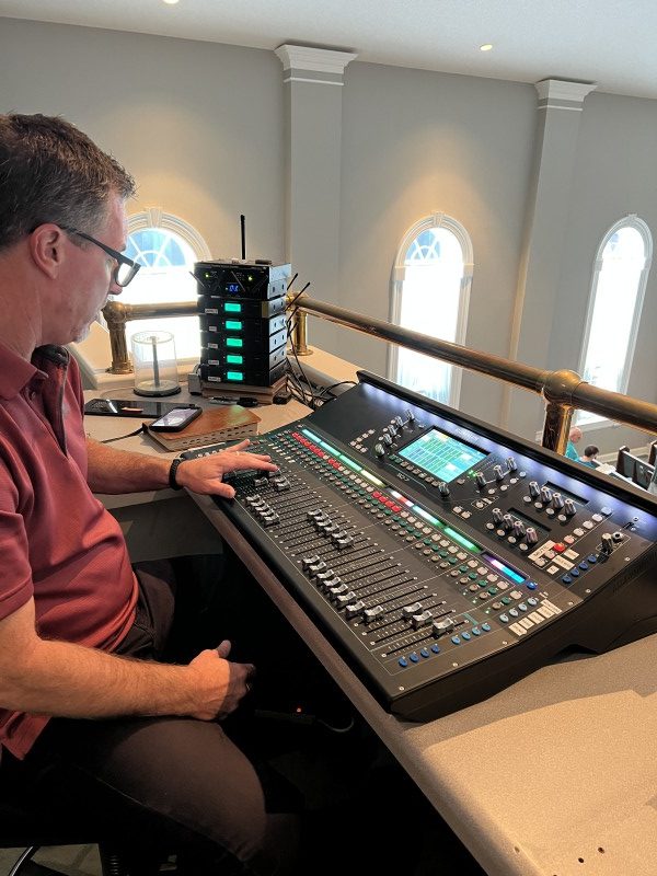 A sound board operator sits at a digital mixer and changes the channels to control audio quality during a live worship event.