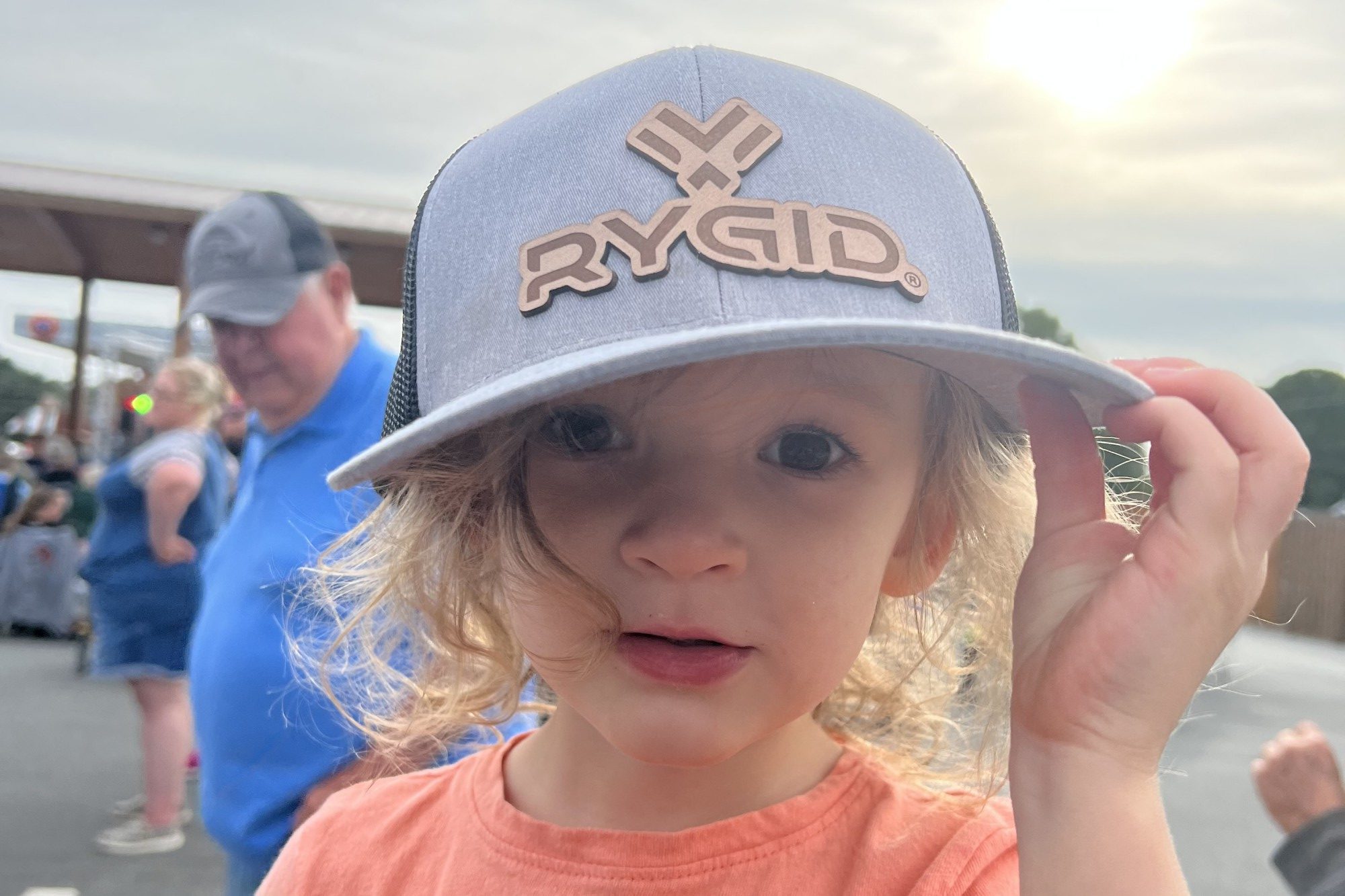 A close-up of an adorable three-year-old girl staring at the camera, holding the brim of a RYGID AV-branded baseball cap.