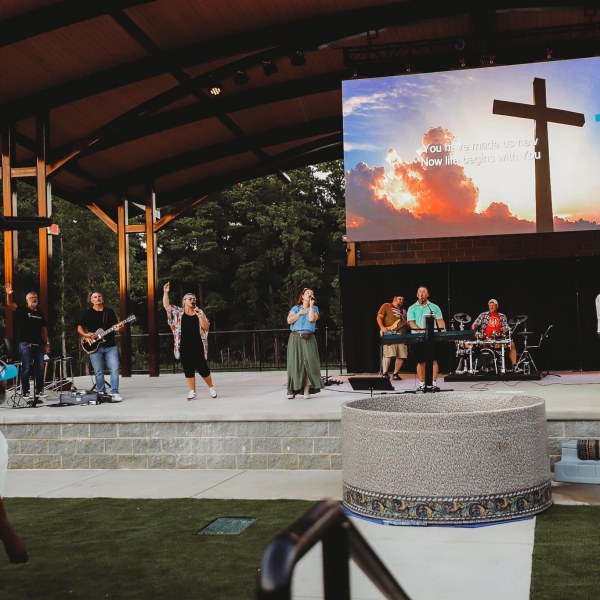Seven people sing praise songs on an outdoor stage covered by a shelter covering. A large screen with a cross and the words to a song are displayed prominently.