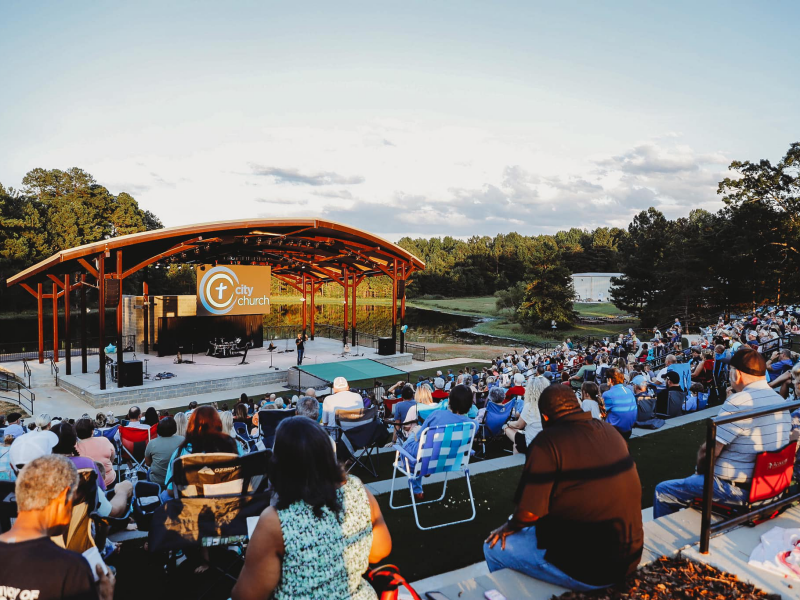 A picture of an amphitheater with a beautiful lake behind it. A crowd of people sit in folding chairs and on concrete steps facing the stage, listening to a performer; the church sound system lets everyone hear her clearly.
