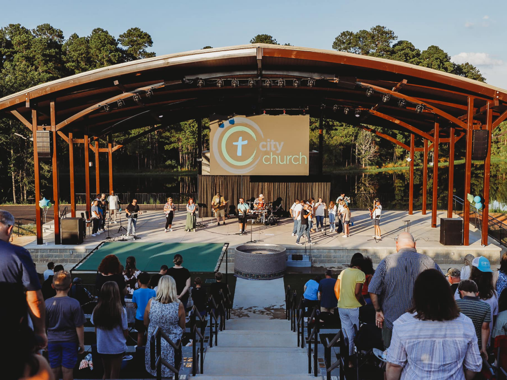 A full amphitheater for a church during a worship service; a large screen displays the church's name and speakers are mounted to get sound to all attendees sitting on the steps.