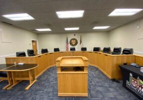 The podium and ten empty seats of city council chambers, all with microphones, recording, and broadcasting equipment.