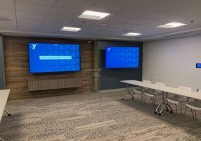 Two large, blue-screened televisions are mounted on the wall of a long conference room. Patterned carpets and long tables with plastic chairs run along the sides of the room. A control panel for the TVs is on the wall.