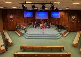 The image is looking down on a church sanctuary, with empty wooden pews and a raised dais with a band and speaker checking the new sound system. Three large video displays hang and show the words of the hymn for congregation members to sing along.