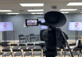 A video camera is in focus in the center of the image, its viewfinder open and showing the stage and empty chairs for the upcoming event. Empty audience chairs and blank screens wait to connect the in-person and virtual attendees to the event.