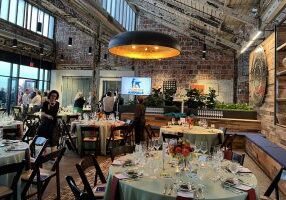 Several round tables are set with elegant place settings in an open venue with brick walls and exposed wooden beams. A video monitor in the back displays "Stand For Animals," the group hosting the fundraiser.