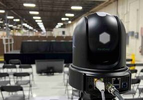 The back of a BirdDog brand webcam and the streaming kit is in focus, with a stage and empty seats out of focus around it.